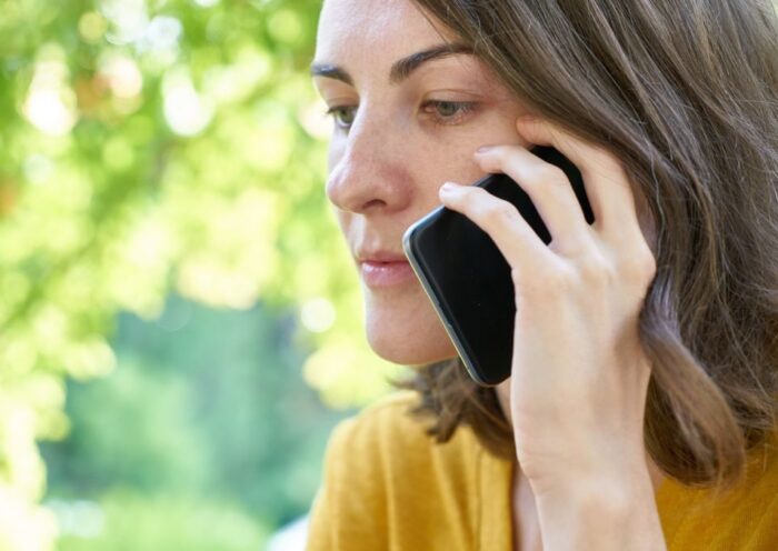 Side profile of a woman using a smartphone outdoors in a sunlit park, wearing a yellow top