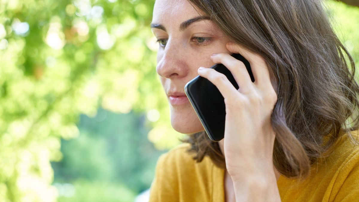 Side profile of a woman using a smartphone outdoors in a sunlit park, wearing a yellow top