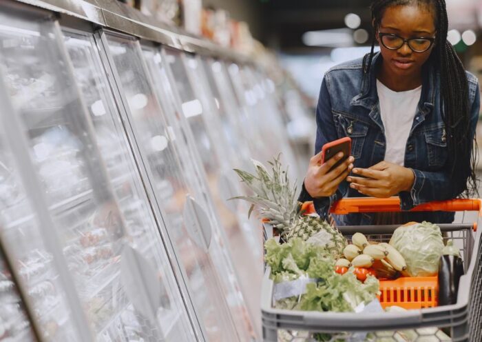 Woman in a grocery store using smartphone while pushing a shopping cart filled with groceries