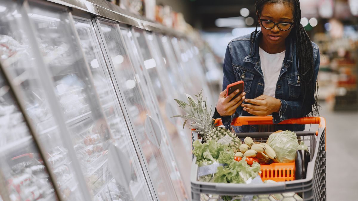 Woman in a grocery store using smartphone while pushing a shopping cart filled with groceries
